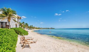 A beach with clear blue water, a row of wooden lounge chairs, and a white building with a thatched roof. - Nikte Ha Unit 2: 2 BR, 2 BA CONDO in Akumal, Sleeps 5 (Akumal)