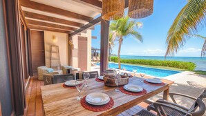 Mesa de comedor de madera dispuesta con platos y copas de vino, con vista a una piscina y al océano.