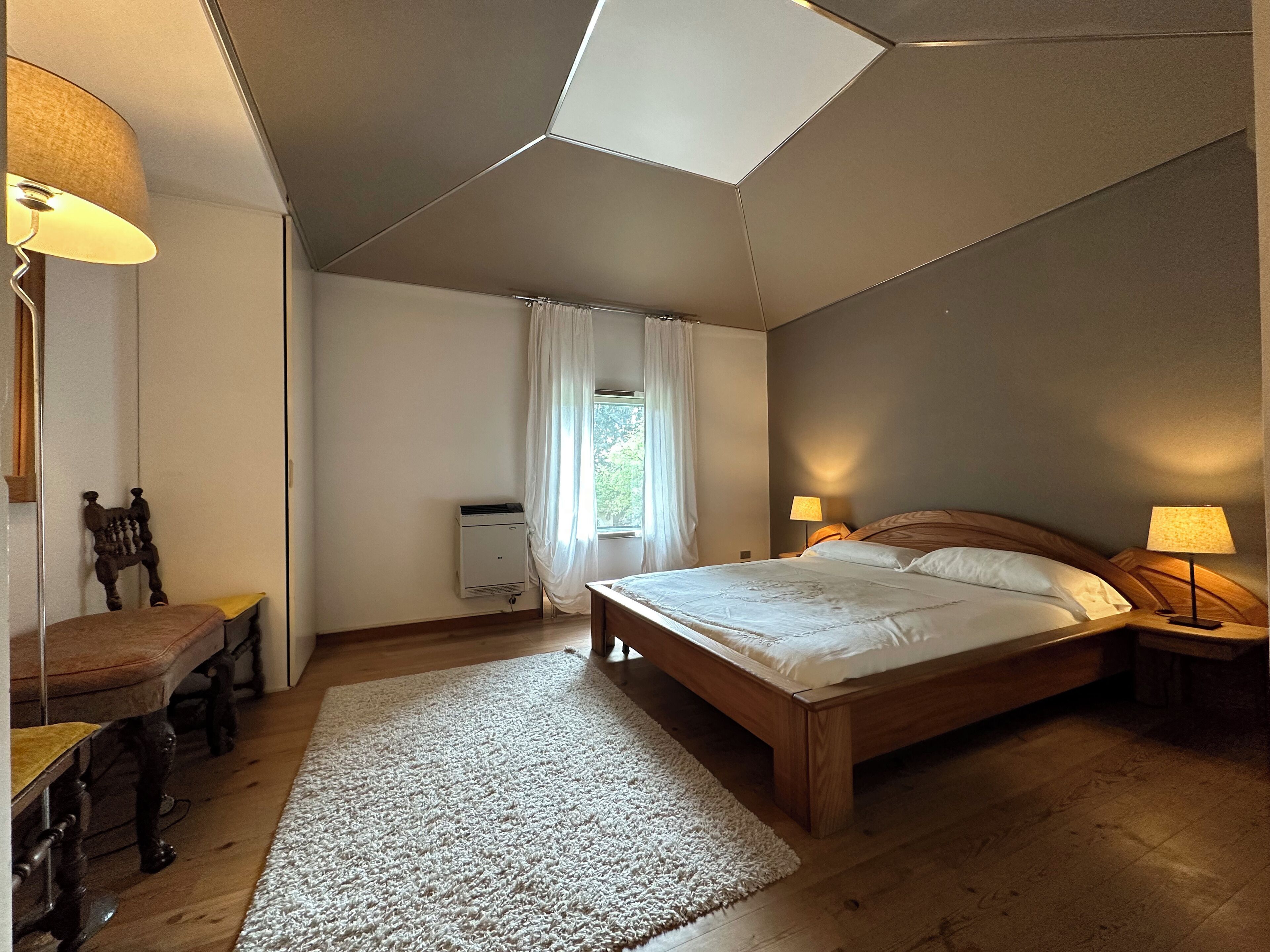Een slaapkamer met een groot bed, twee nachtkastjes met lampen, een houten stoel, een kleine tafel, een radiator en een raam met gordijnen.