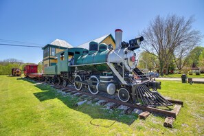 Una locomotora de vapor verde sobre una vía con un vagón de freno y un coche rojo, rodeado de hierba y árboles.