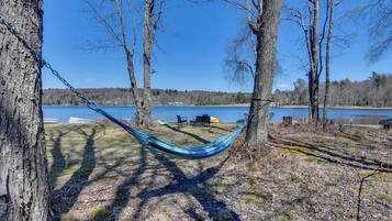 A hammock between trees overlooking a lake with chairs and a table nearby.