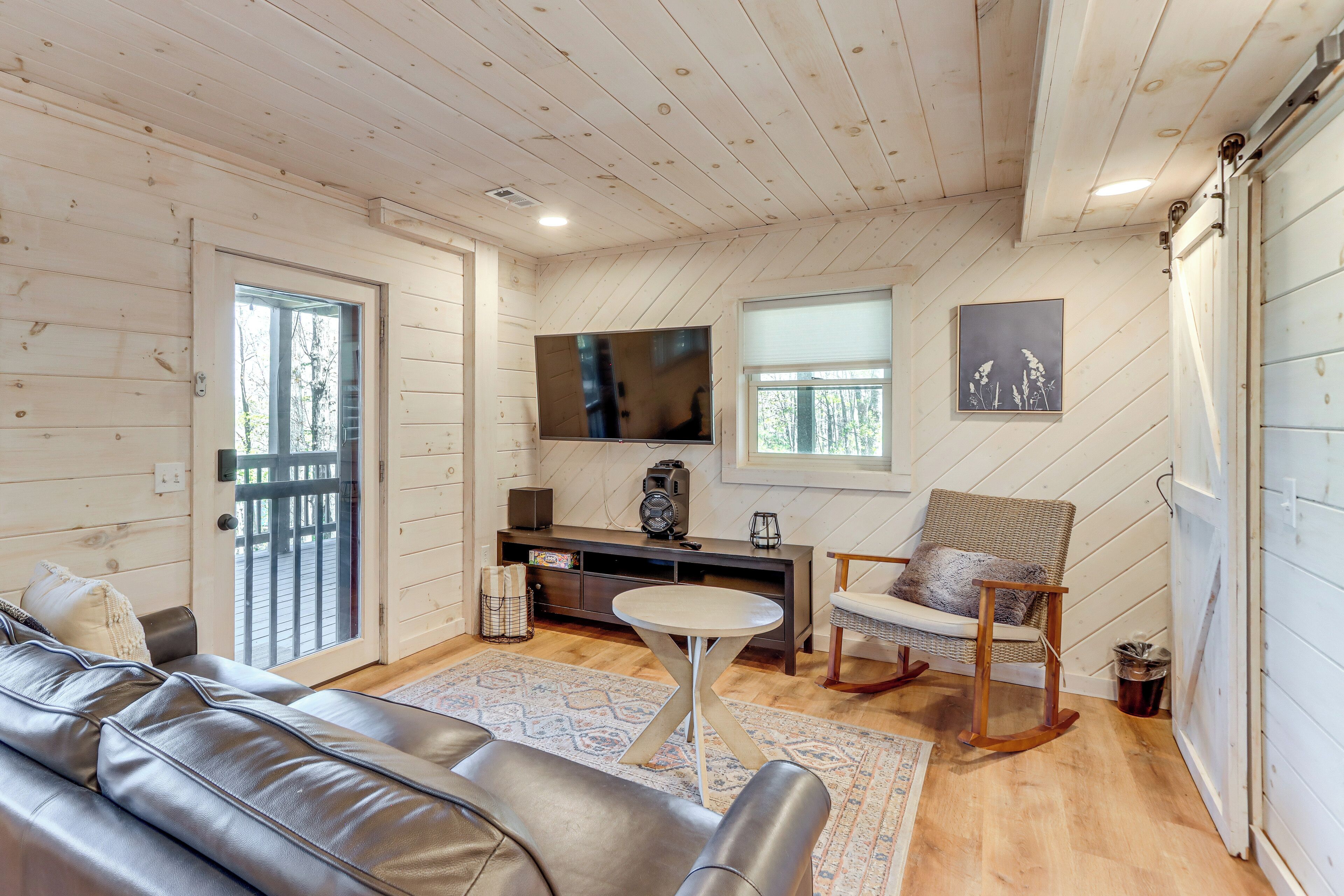 A cozy living room with a leather sofa, a round coffee table, a wooden chair, a TV mounted on the wall, and a sliding door leading to an outdoor area.