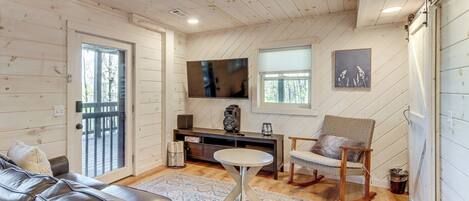 A cozy living room with a leather sofa, a round coffee table, a wooden chair, a TV mounted on the wall, and a sliding door leading to an outdoor area.