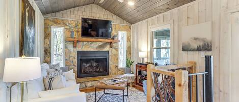 A cozy living room with a stone fireplace, a television, a wooden ceiling fan, and a large window.