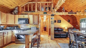 A rustic cabin interior with wooden floors, a kitchen with a large island, a dining area with a wooden table and chairs, and a fireplace. - Waterfront Anglers Oasis in The Maine Highlands! (Carmel)