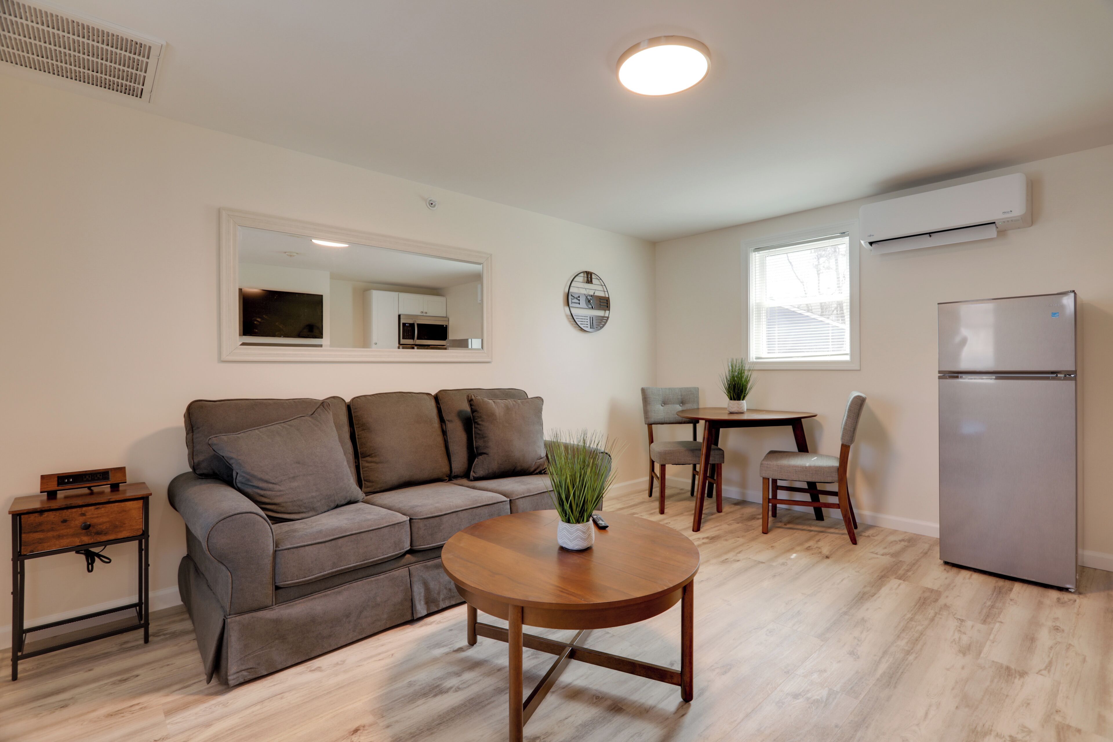 A modern living room with a grey sofa, a wooden coffee table, a dining table with chairs, and a built-in air conditioner.