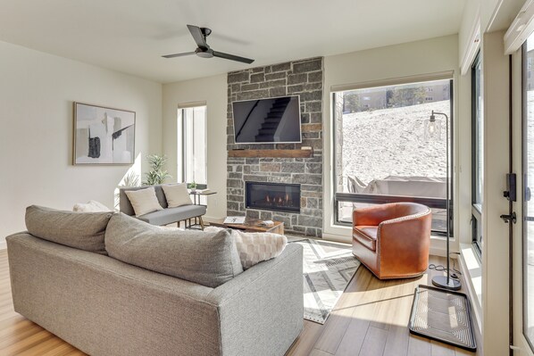 A modern living room with a stone fireplace, a sofa, a leather armchair, and a large window with a view of a snowy landscape. - Walk to Ski Lift: Granby Ranch Family Retreat! (Granby)