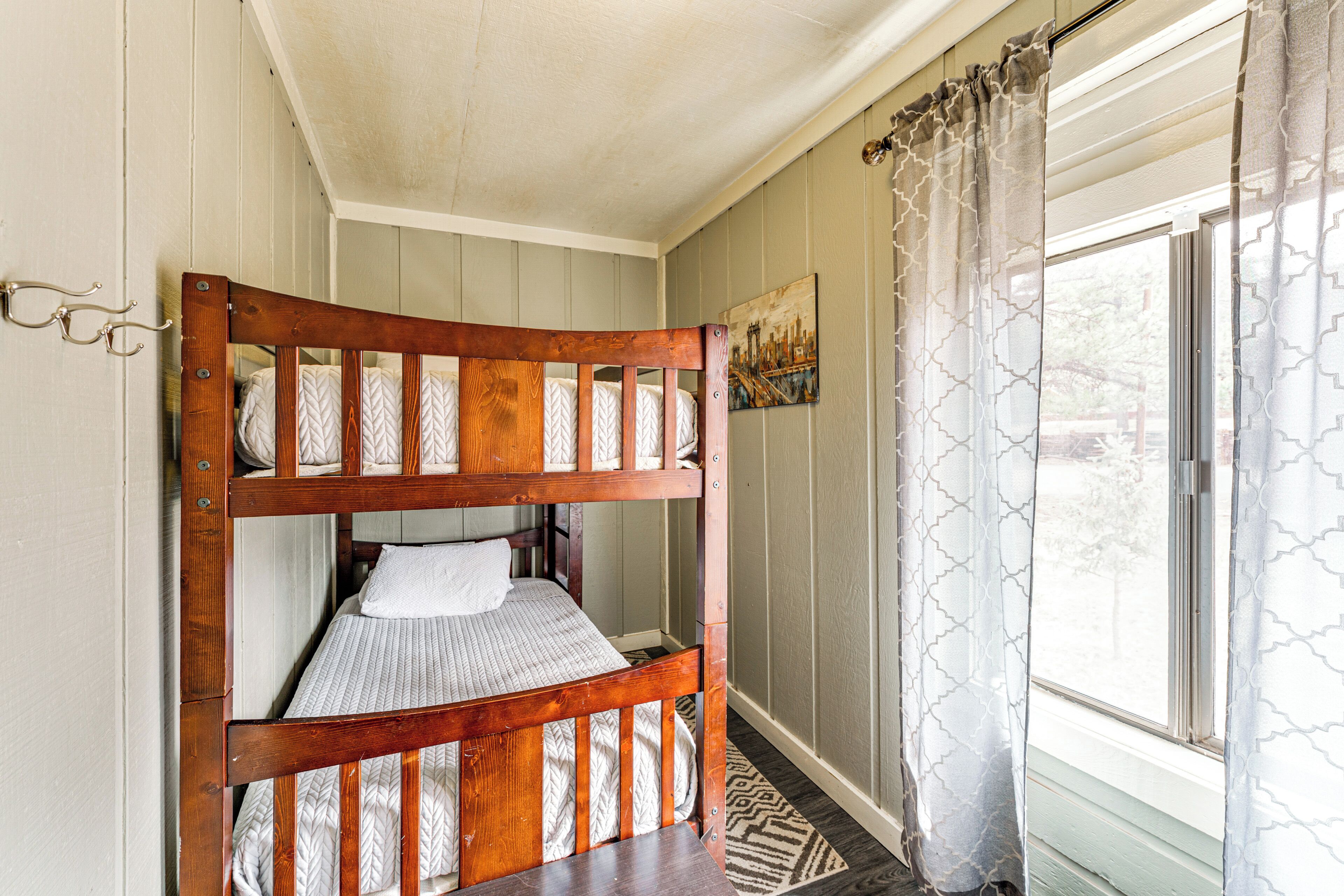 A bunk bed with a wooden frame and patterned bedding, a window with patterned curtains, and a painting on the wall.