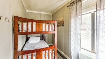A bunk bed with a wooden frame and patterned bedding, a window with patterned curtains, and a painting on the wall.