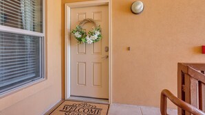 Una puerta color beige con un adorno de bienvenida, un felpudo y una lámpara de pared.
