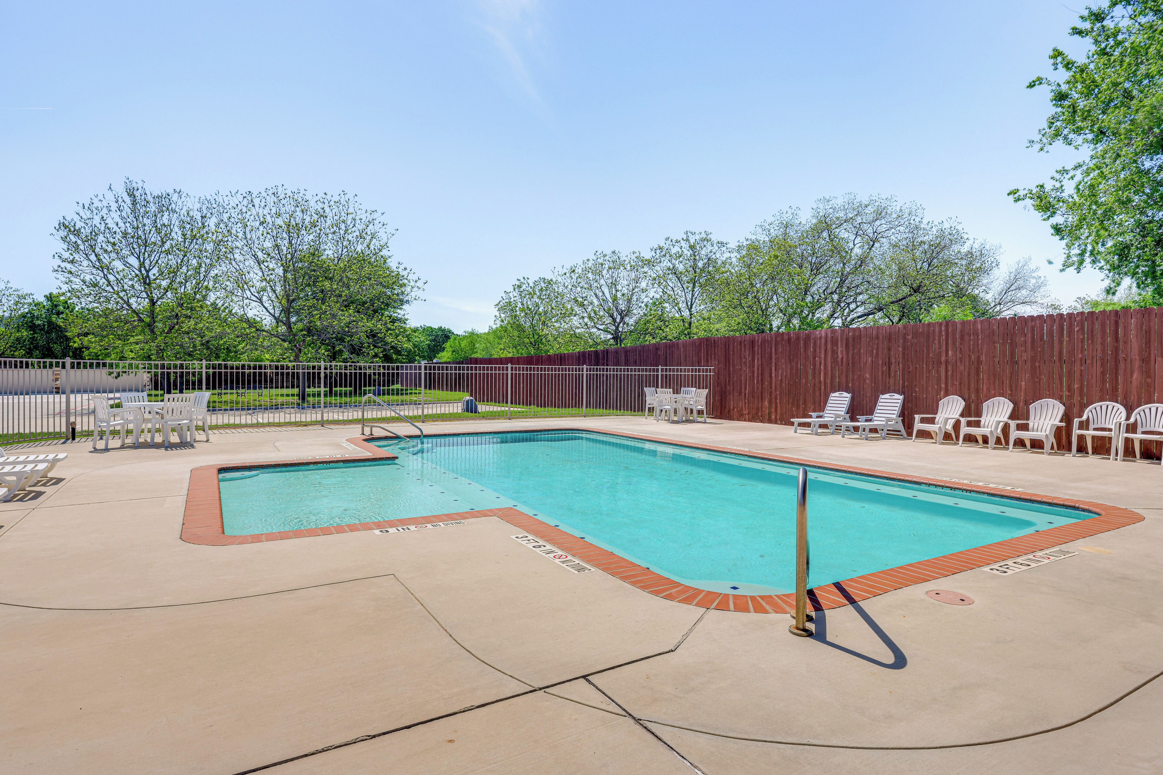 A rectangular swimming pool with a wooden deck surrounding it, a metal fence, and white lounge chairs.