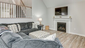 A living room with a grey sectional sofa, a fireplace, a TV mounted above it, and a staircase with white railings.