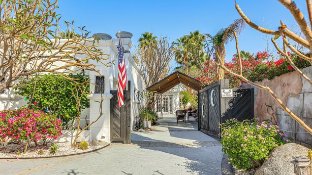A paved pathway leading to a building with a flag, surrounded by lush greenery and flowering plants.