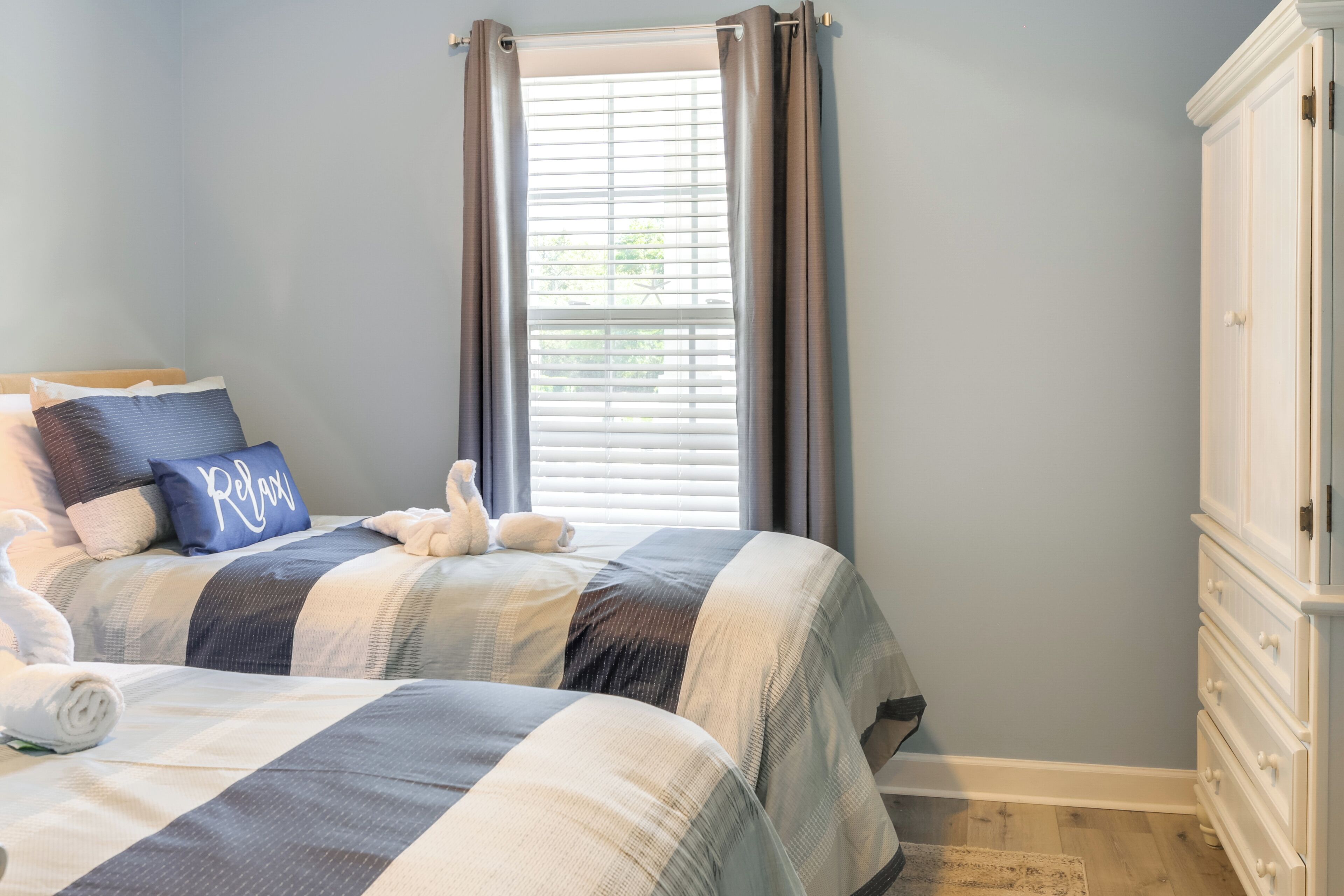 A bedroom with two beds, a dresser, a window with curtains, and a blue pillow with the word "Relax".