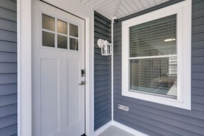 L’extérieur d’une maison avec une porte blanche, une fenêtre équipée de stores et un petit panneau indiquant une surveillance 24 heures sur 24.