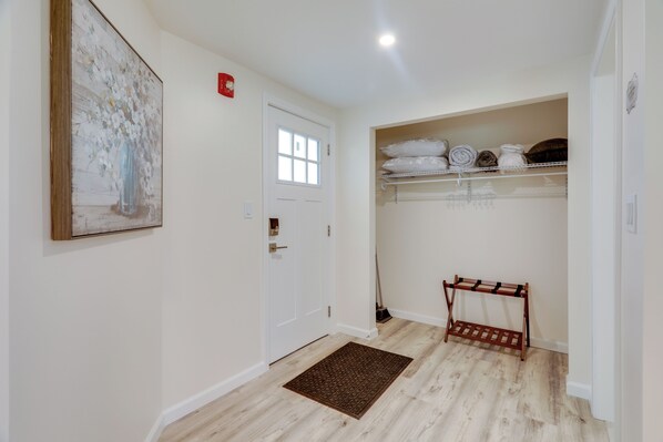 Un couloir avec une porte blanche, un plancher en bois, un paillasson et une étagère où sont pliées des serviettes.