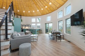 Enjoy a spacious open-plan living area with a high ceiling and large windows, a comfortable seating area, dining table, and kitchen with bar seating.