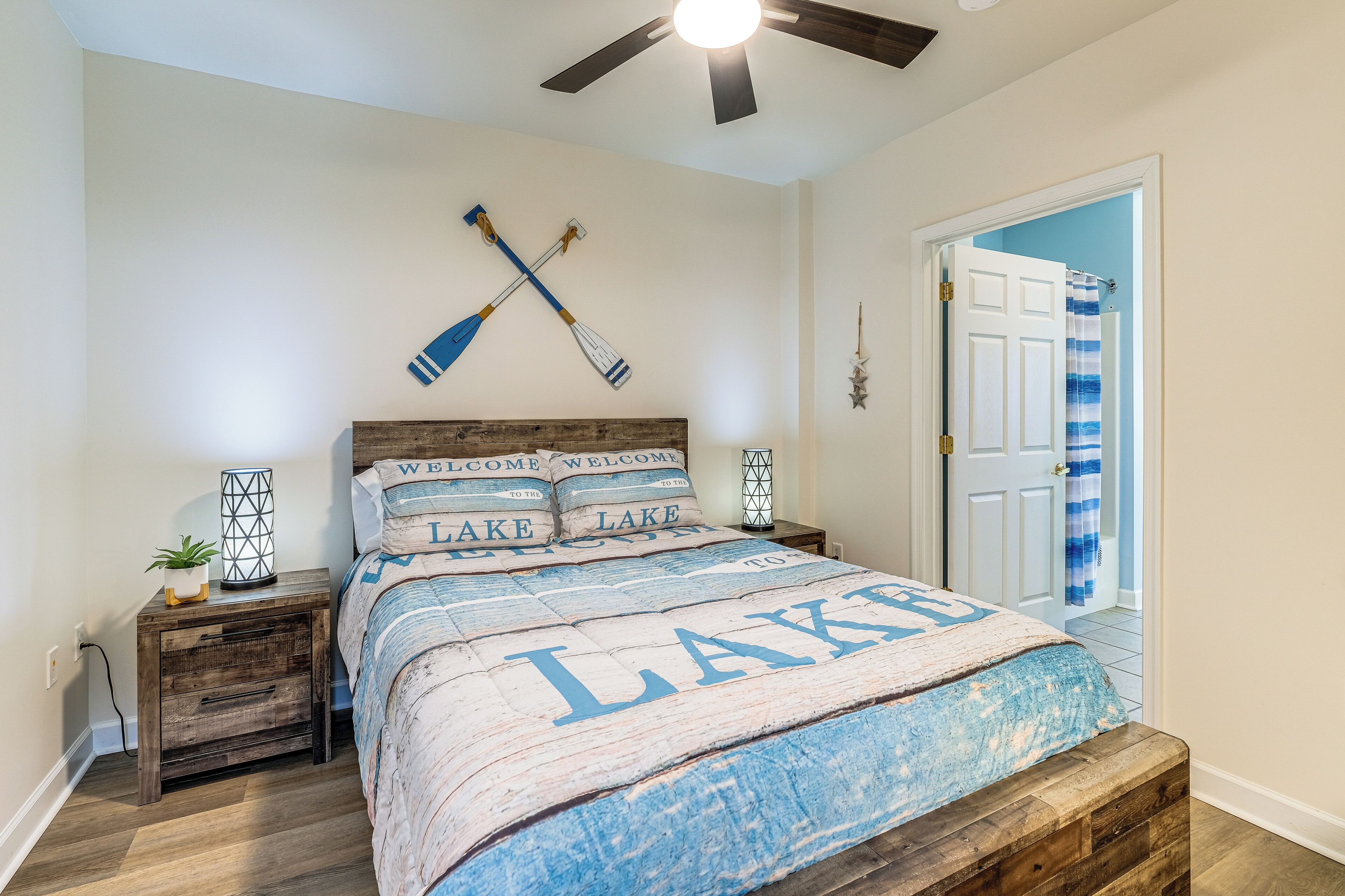 A bedroom with a bed featuring a quilt with 'LAKE' text, wooden headboard, two bedside tables with lamps, and a ceiling fan.