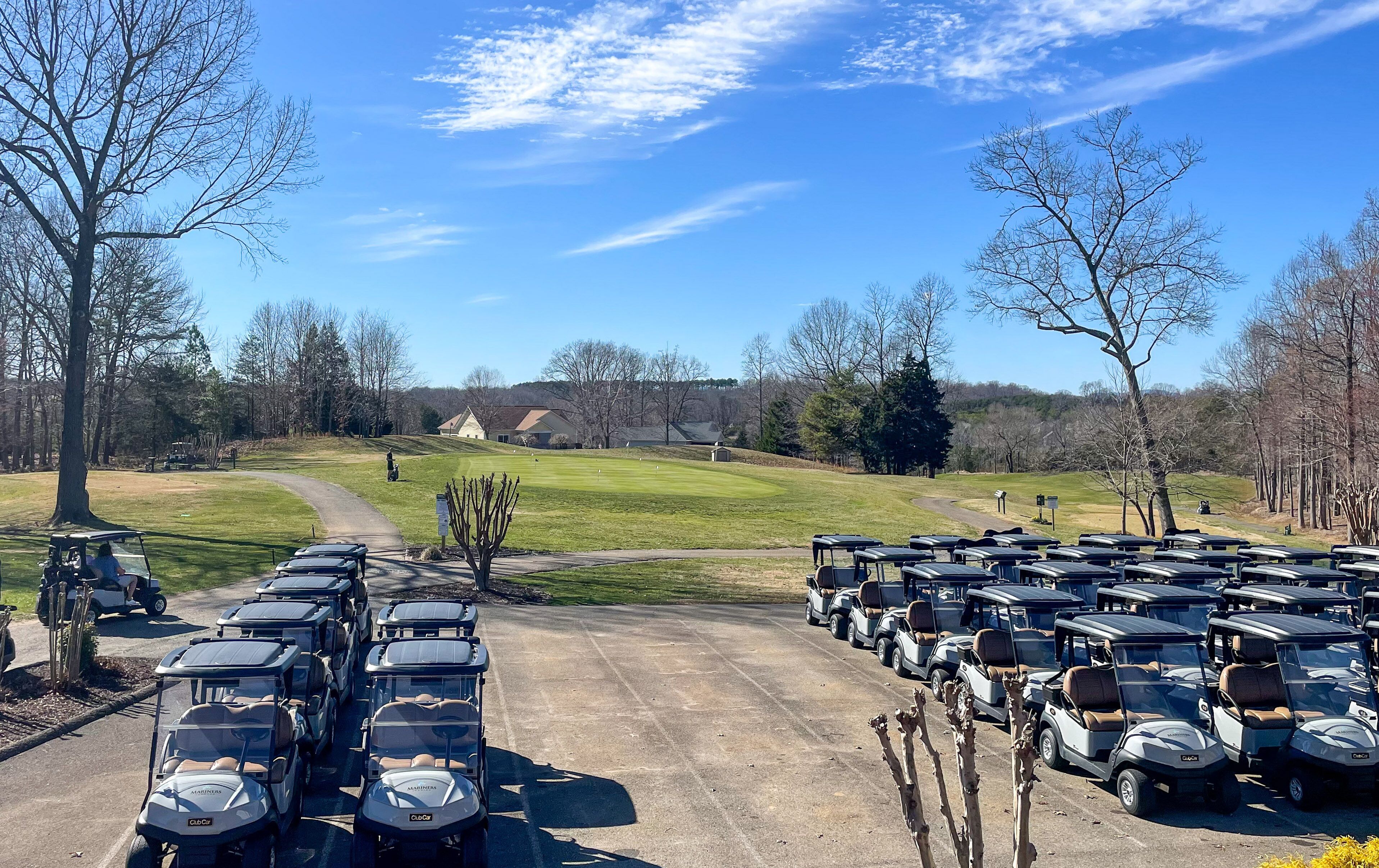 A parking lot with golf carts lined up, a golf course in the background, and a clear sky.