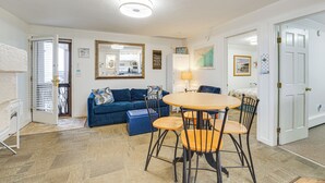Ein Wohnzimmer mit blauem Sofa, einem Holzesstisch mit vier Stühlen, einer Küche im Hintergrund und einer weißen Tür.