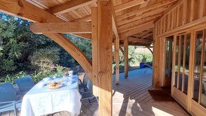 Een houten terras met een ontbijttafel, omringd door stoelen, en een overdekt gedeelte met grote ramen.