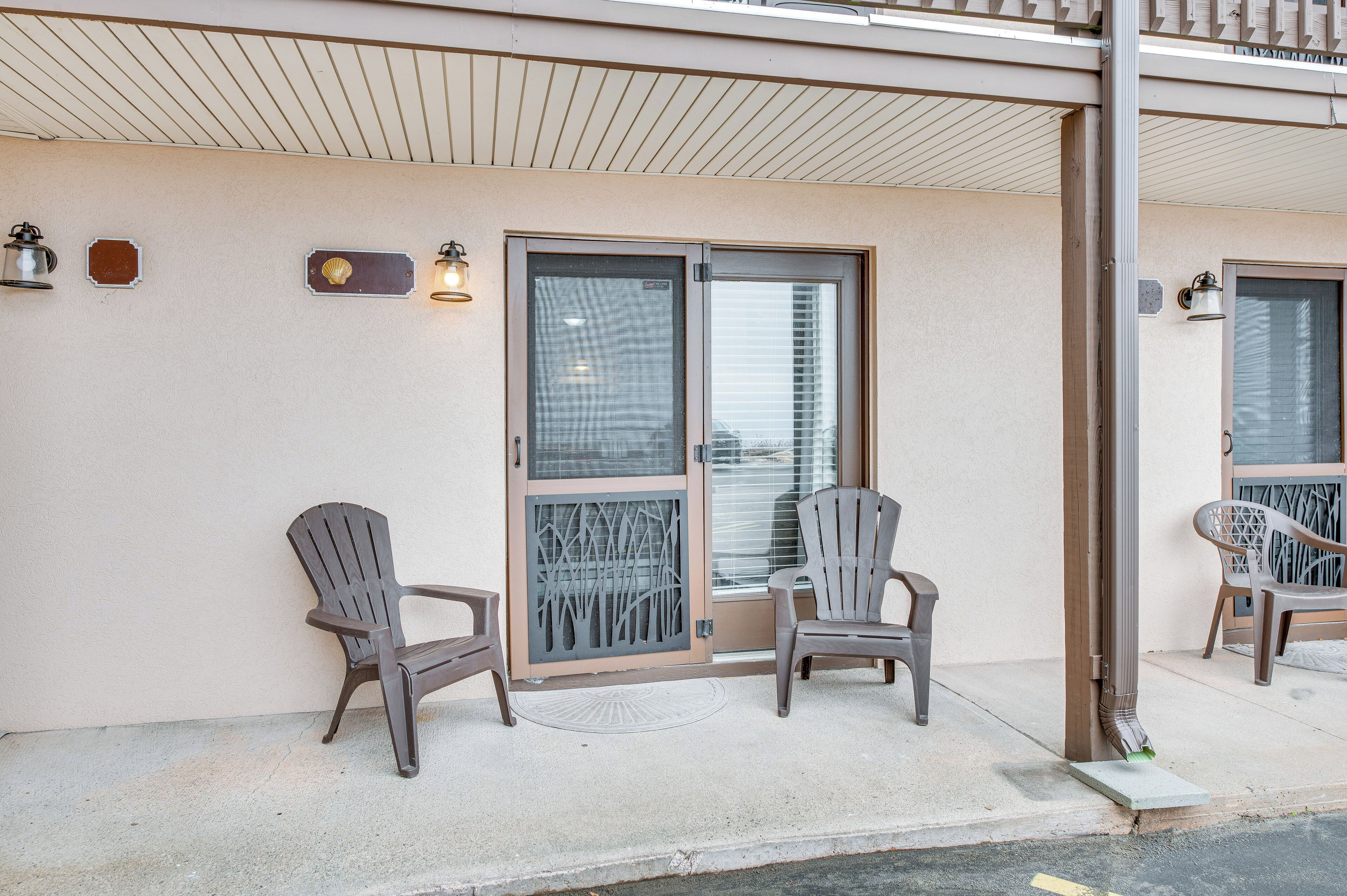 A building entrance with two Adirondack chairs outside, a glass door, and a small sign on the wall.