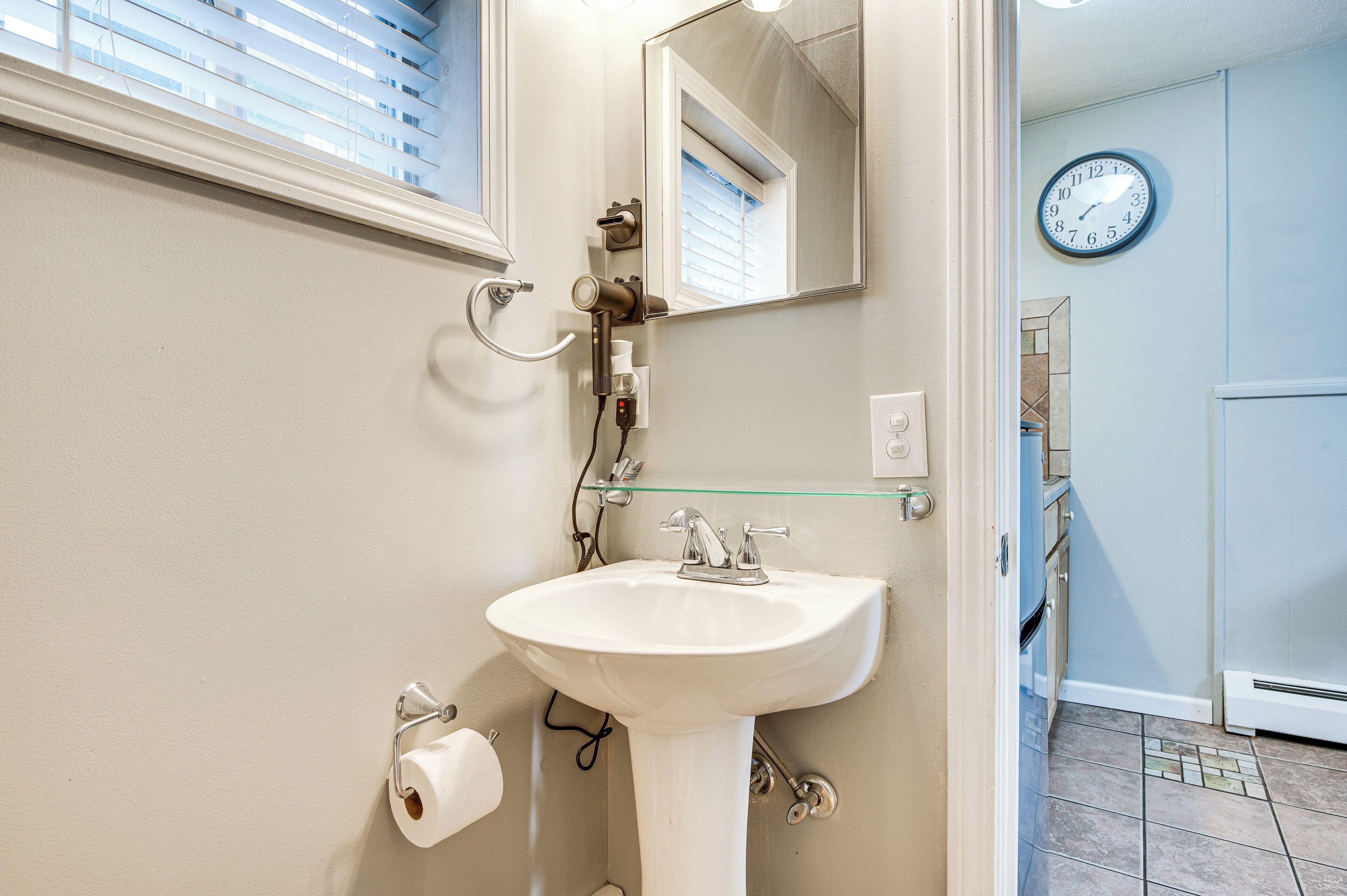 Une salle de bain avec un lavabo blanc, un miroir, une horloge au mur et une fenêtre avec des stores.