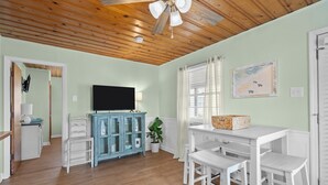 Una habitación con techo de madera, un televisor sobre un mueble, una mesa de comedor con sillas y una ventana con cortinas.