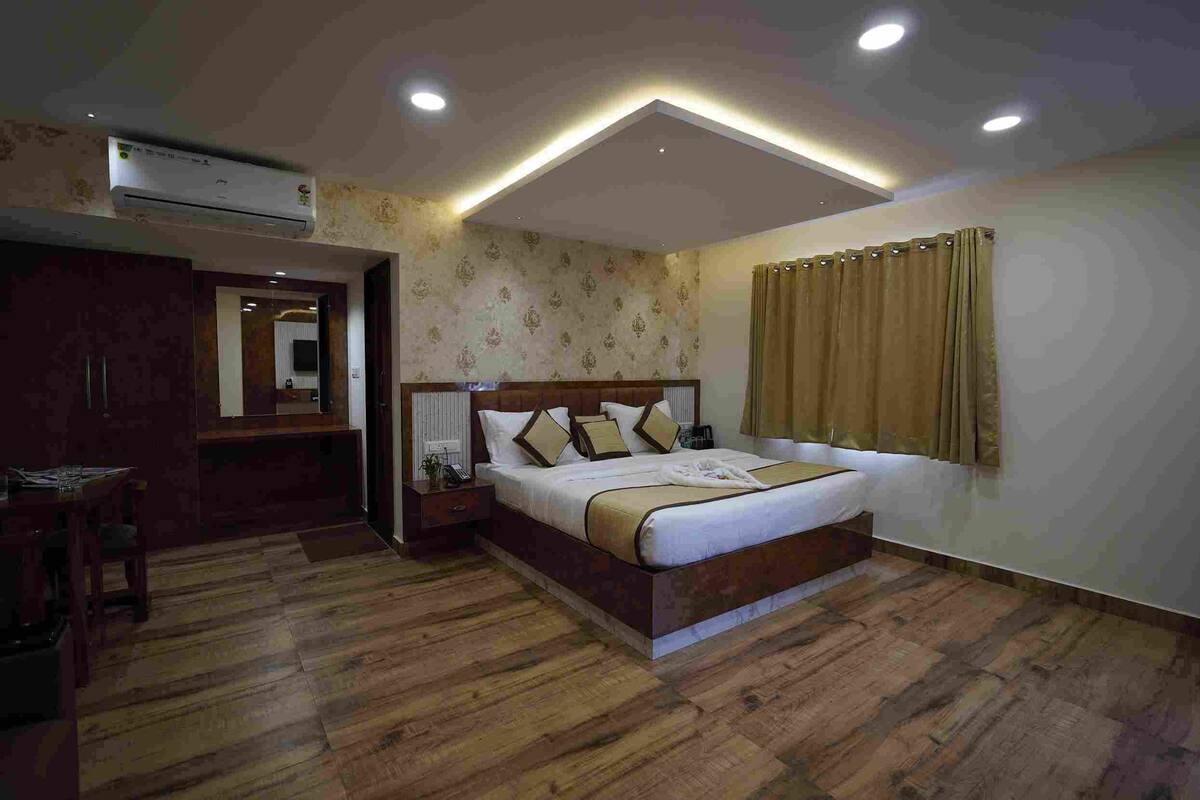 A bedroom with a wooden floor, a bed with a headboard, a bedside table, a nightstand, an air conditioning unit, and a window with curtains.