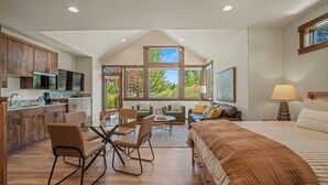 A spacious room with a large bed, a dining table with chairs, a kitchen area, and a view of trees through large windows.