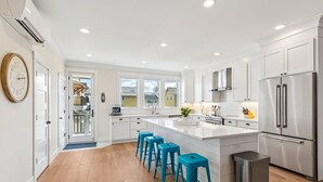 A modern kitchen with white cabinetry, a central island with blue bar stools, stainless steel appliances, and a large window providing natural light. - Seabrook King Tide | Oceanfront, Hot Tub, Pets 4BR | Sleeps 8, Pacific Landing (Pacific Beach)