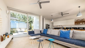 Een moderne woonkamer met een modulaire bank, een glazen deur naar een terras en een keuken met witte kasten en een plafondventilator.