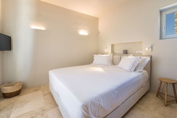 A neatly arranged bedroom with a large bed, a flat-screen TV mounted on the wall, and a small wooden stool. - Aequora Villa II - Beachfront, 5 min away from the Golden Beach, Infinity Pool (Paros, Cyclades)