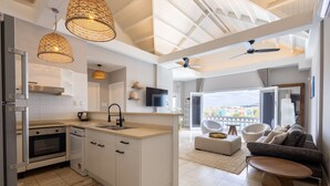 A modern kitchen with white cabinetry, a built-in oven, and a large island with a sink. There is a ceiling fan, a TV mounted on the wall, and a view of the city through large windows. - Chic City View Residence by the Bay (Willemstad)