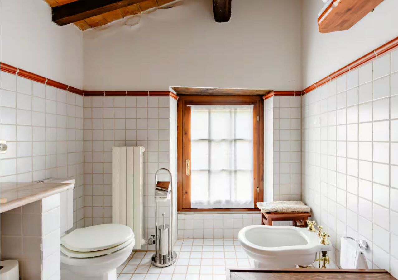 Ein Badezimmer mit Toilette, Bidet und Badewanne, ein Fenster mit Vorhängen und einer Holzbalkendecke.