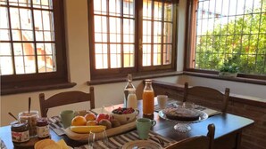 Une table de petit-déjeuner dressée avec du pain, des fruits et des boissons, et offrant une vue sur la verdure à travers de grandes fenêtres.