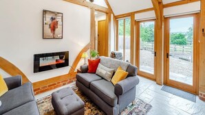 Un salon spacieux avec un canapé gris, un téléviseur à écran plat, des poutres en bois et de grandes fenêtres.