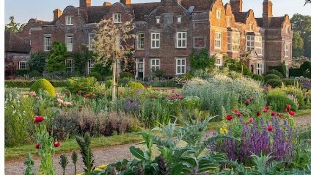 Un grand bâtiment en briques, doté de nombreuses cheminées, entouré d’un jardin bien entretenu où poussent diverses plantes et fleurs.