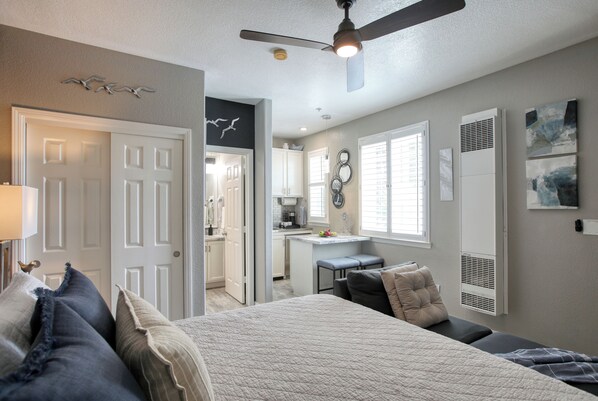 A bedroom with a bed, pillows, a ceiling fan, and a view into the adjacent kitchen.