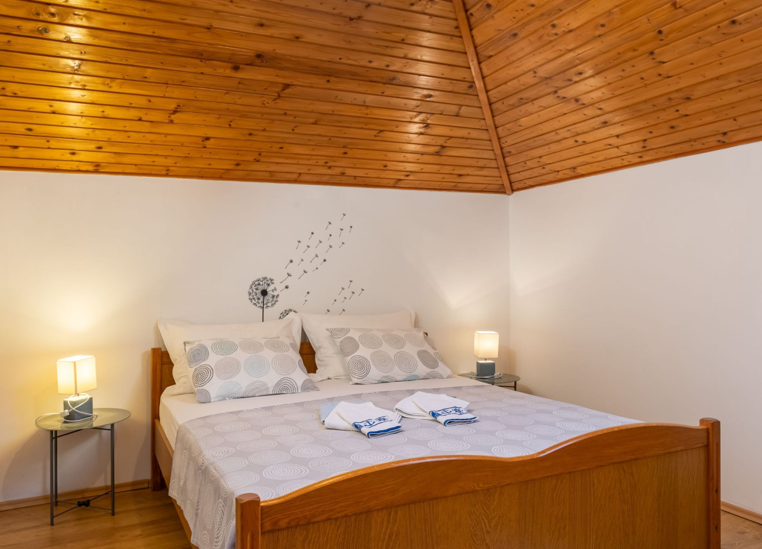 A bedroom with a wooden ceiling, a bed with patterned bedding, two bedside tables with lamps, and a wall decoration of dandelions.