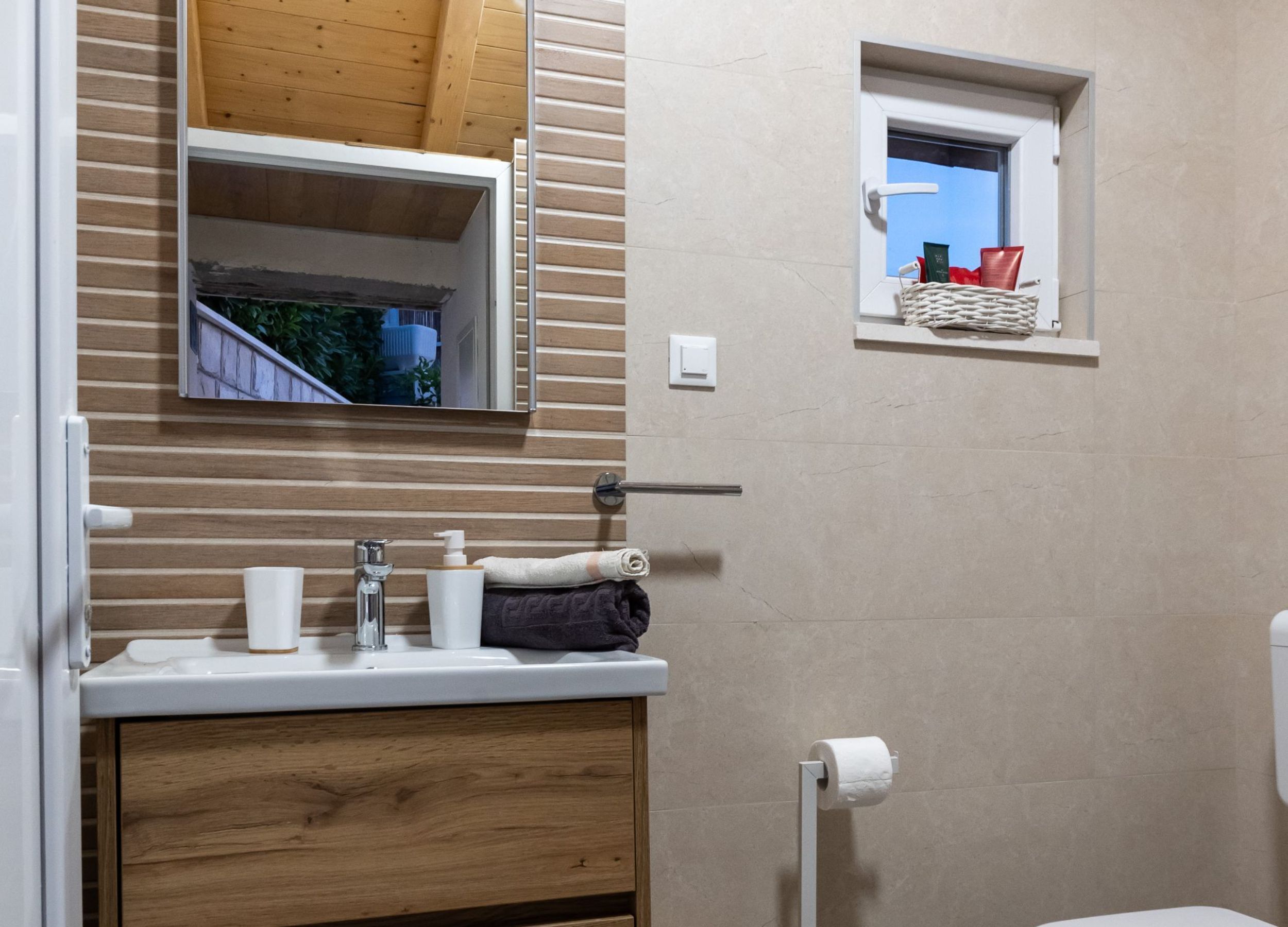A bathroom with a wooden vanity, a mirror, a window with a basket, and a toilet.