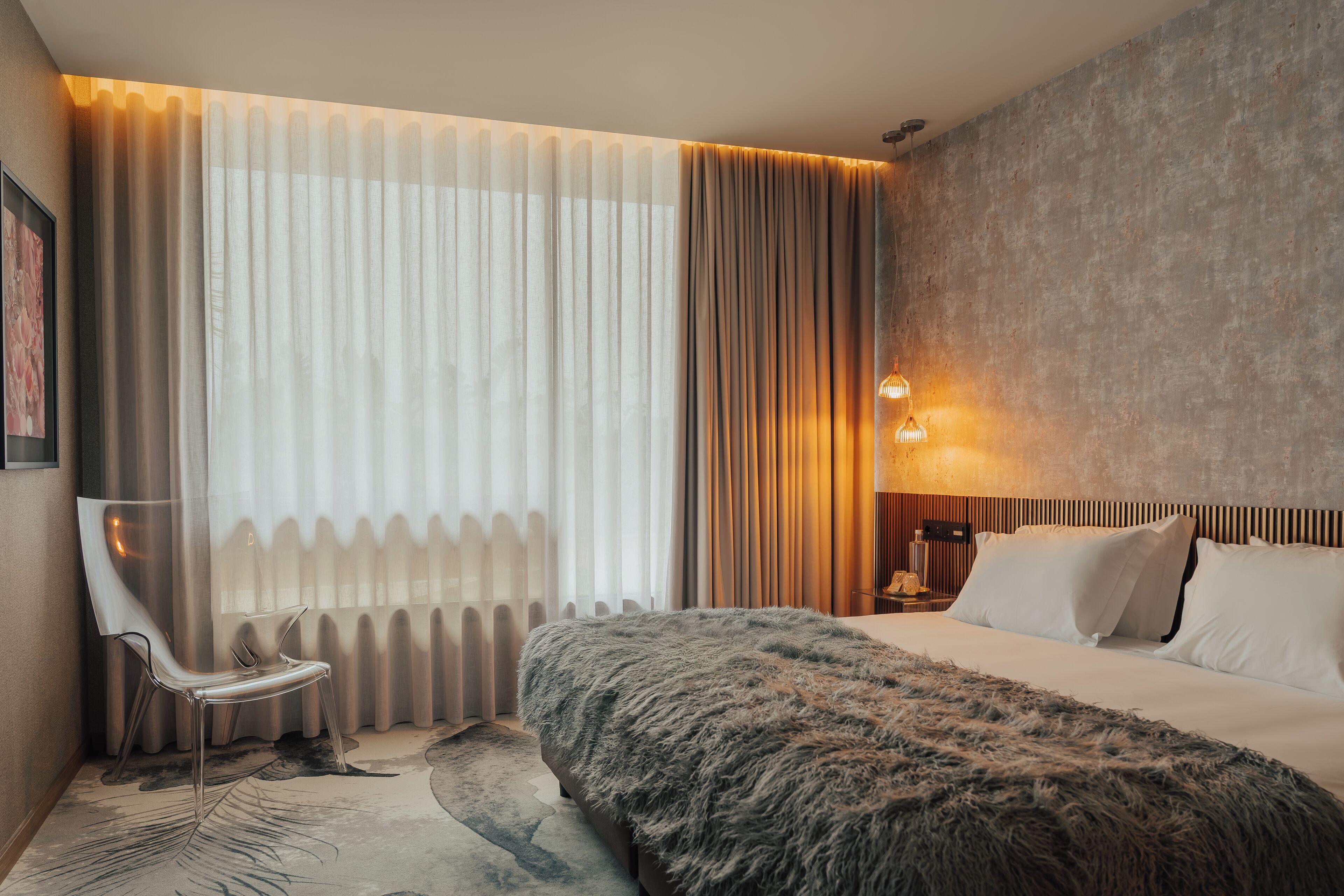 A modern hotel room with a large bed, a chair, a nightstand, and a wall-mounted lamp.
