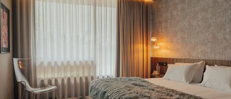 A modern hotel room with a large bed, a chair, a nightstand, and a wall-mounted lamp.