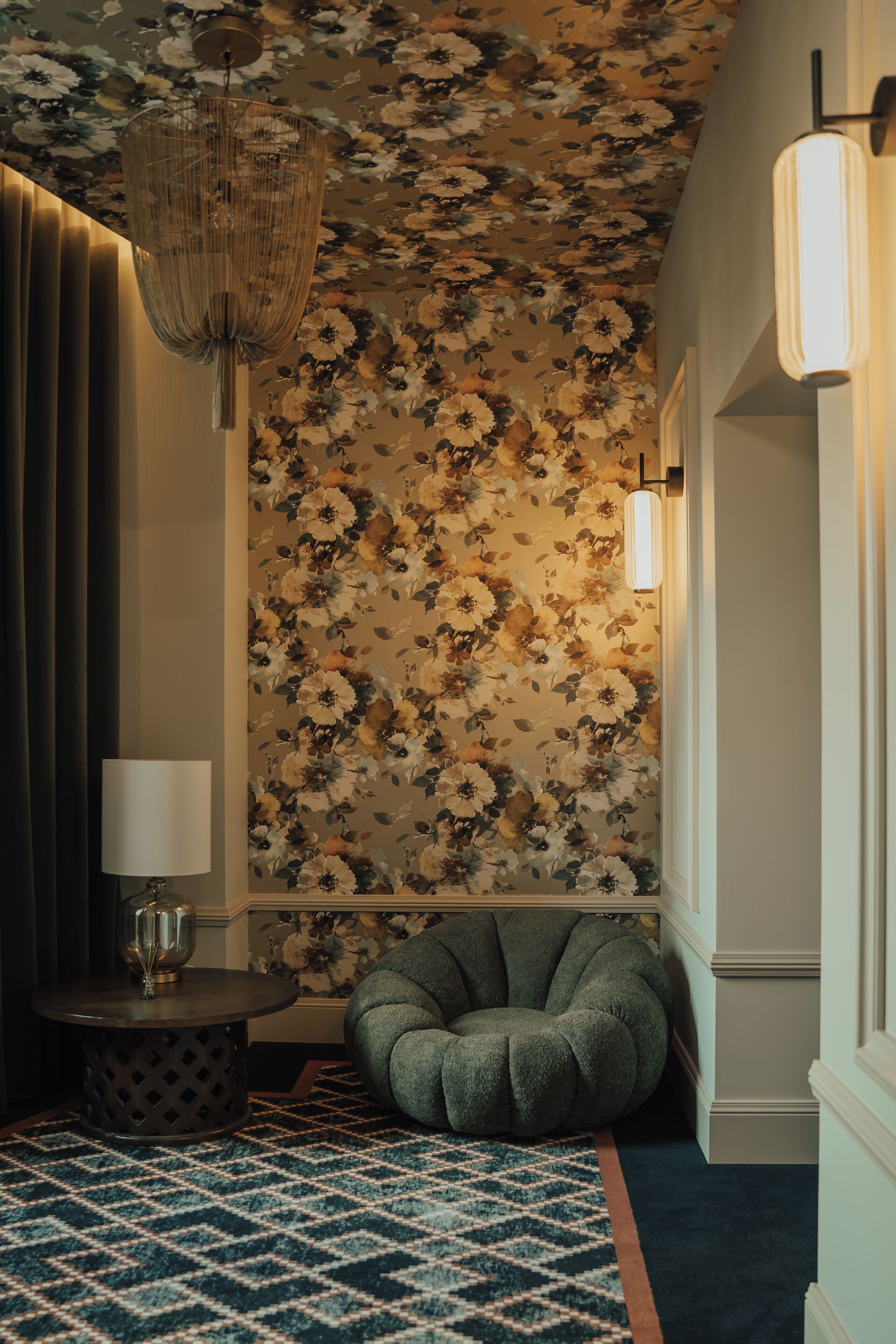 A room with a floral wallpaper, a round table with a lamp, a green bean bag chair, and a patterned rug.