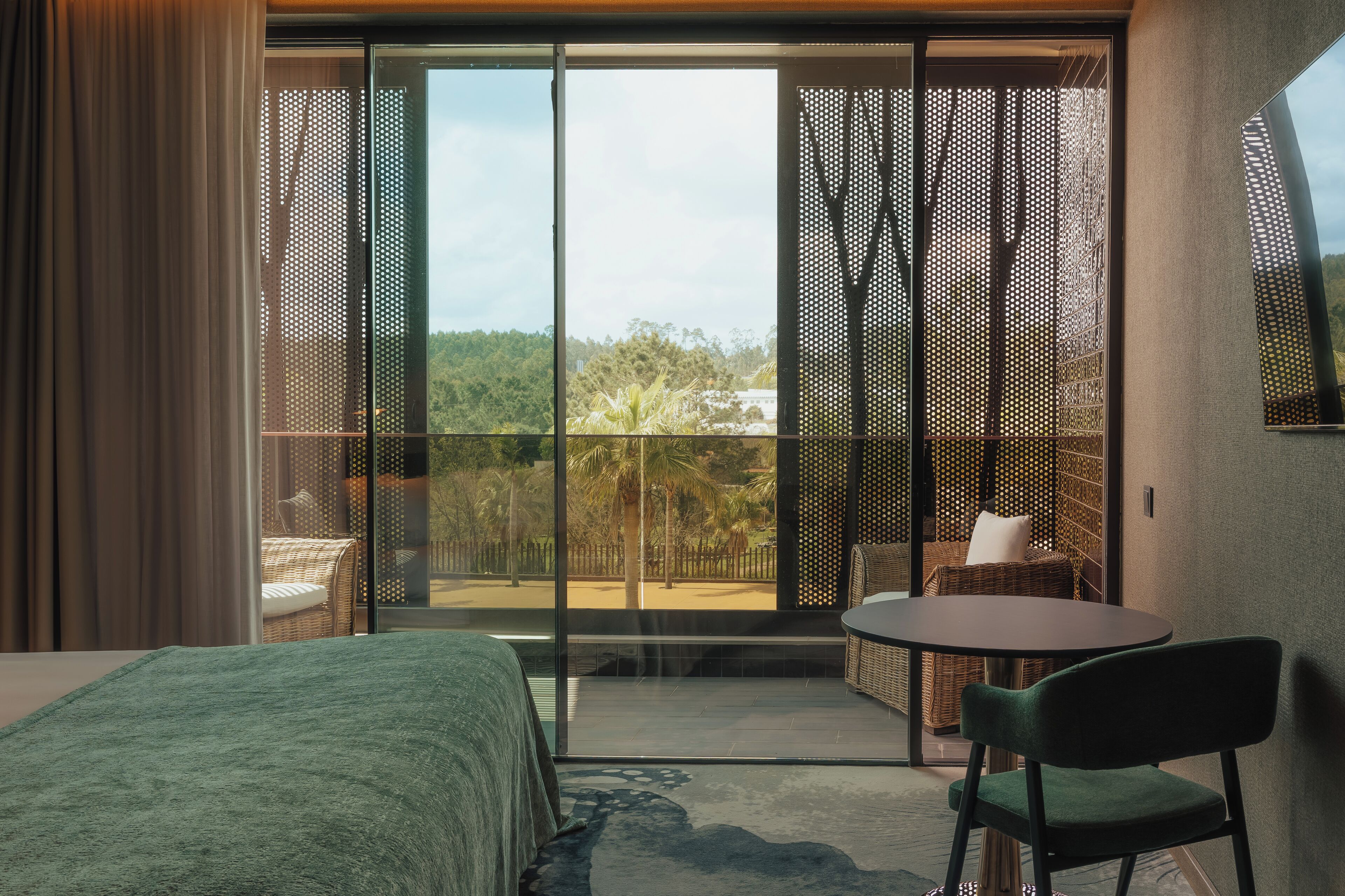 Un dormitorio con una cama, una mesa redonda y una silla. Hay un balcón con vistas a árboles y una valla.