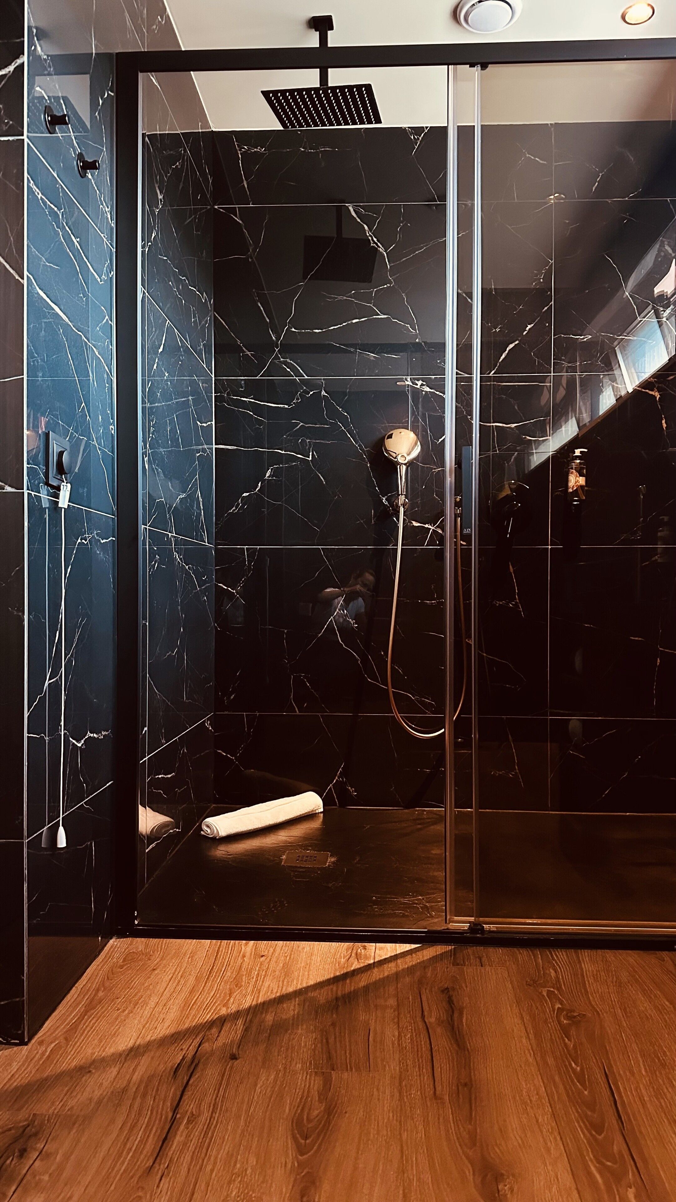 A modern bathroom with a glass shower enclosure, marble walls, and a wooden floor.