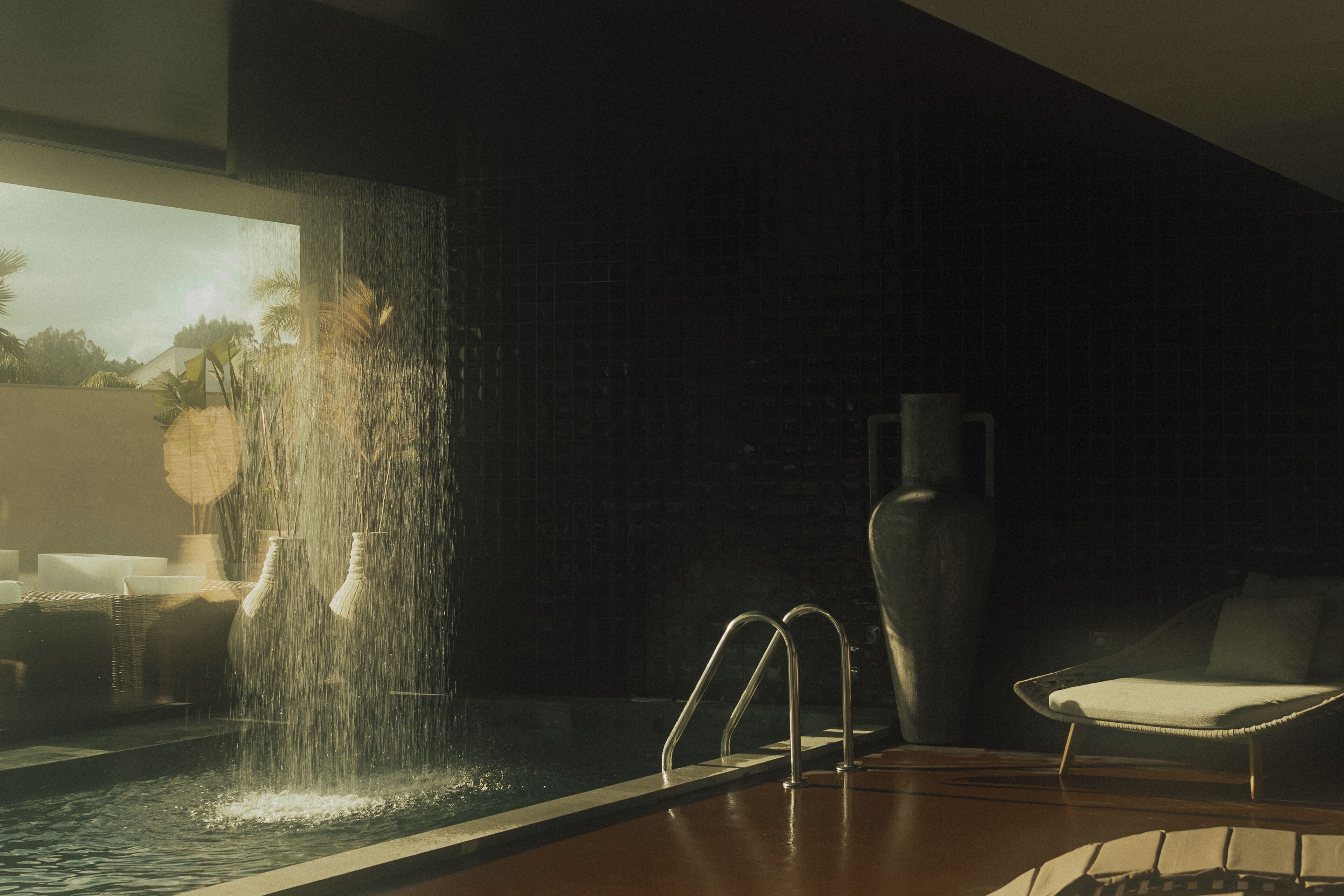 A modern indoor pool area with a waterfall feature, a lounge chair, and a large vase.