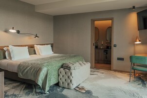 Una habitación de hotel moderna con una cama grande, un banco tapizado en verde, una puerta de madera que da a un baño y una silla verde.