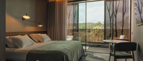 Habitación de hotel con cama, una silla, una mesa, balcón con vistas y una lámpara de pared.