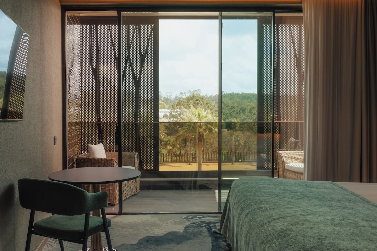 Une chambre moderne avec un grand lit, une petite table et une chaise. Elle dispose d’un balcon avec vue sur les arbres et une clôture.